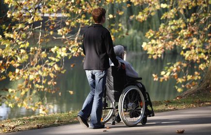 Person schiebt ältere Person im Rollstuhl am Seeufer mit Herbstlaub