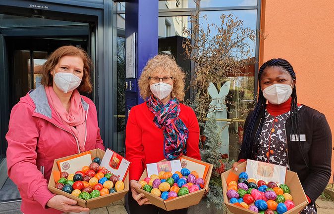Drei Frauen mit FFP2-Masken halten Schalen mit bunten Ostereiern vor einem Gebäude