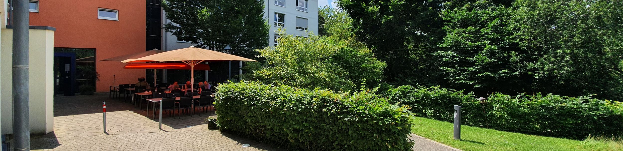 Hofterrasse mit orangem Sonnenschirm, Tischen und sitzenden Personen, umgeben von Bäumen