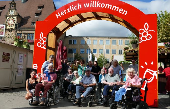Gruppe älterer Menschen in Rollstühlen unter Willkommensbogen des Heilbronner Weindorfs