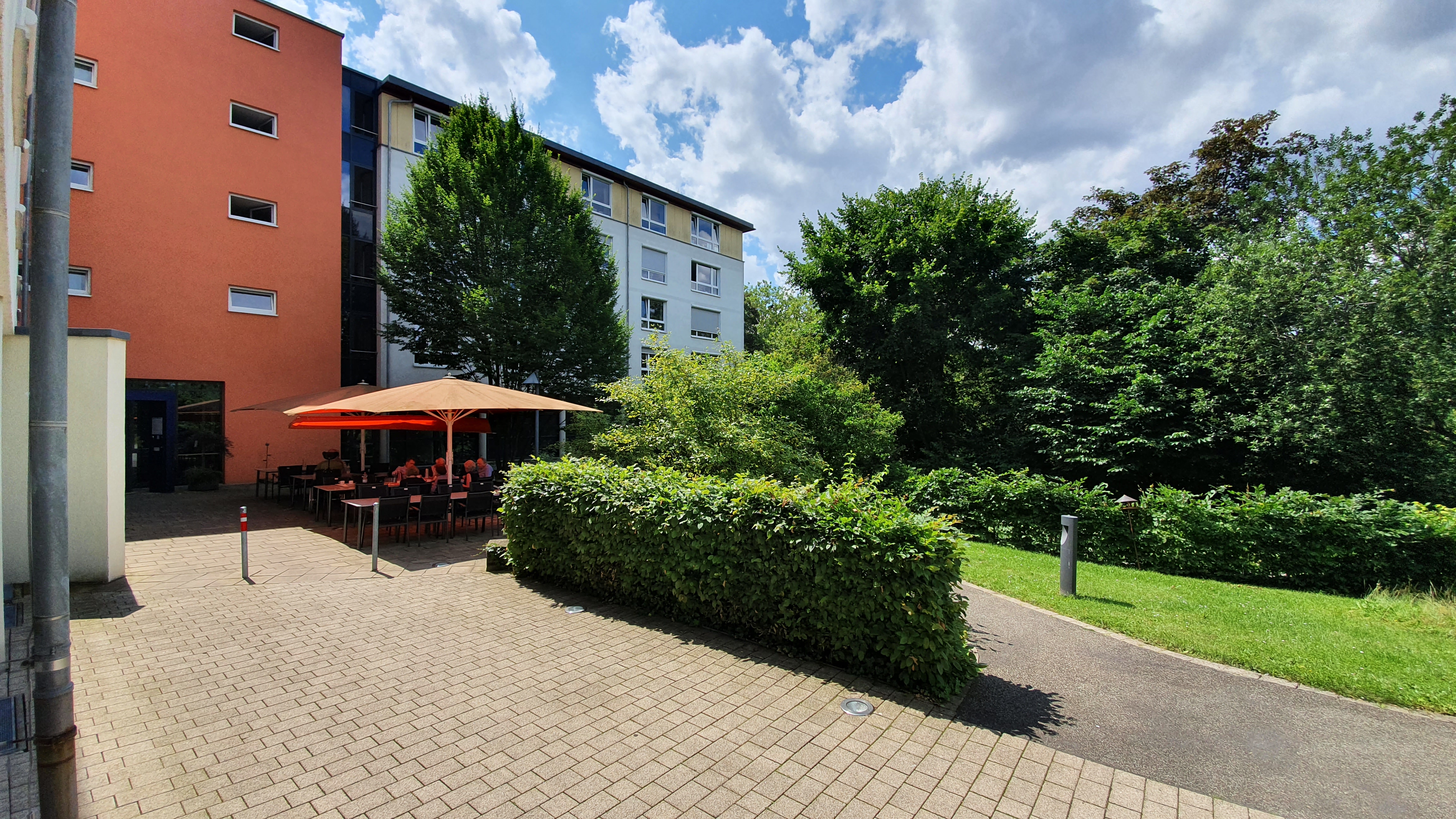Hofterrasse mit orangem Sonnenschirm, Tischen und sitzenden Personen, umgeben von Bäumen